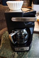 Full view of black DeLonghi drip coffee maker with glass carafe and white coffee filter on top.