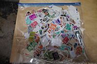 Large sealed plastic zip bag filled with a large quantity of used postage stamps in various colors and designs from many countries spread out inside the bag.