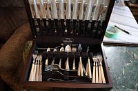 Open wooden case displaying full set of knives in upright slots and forks, spoons and serving utensils below.