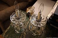 Pair of clear glass mantle lamps with square bases, glass prism pendants, candle-style bulbs, and electrical cords resting on a green marble table.