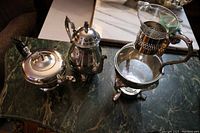 Silver plate tea pot, coffee pot, glass carafe in silver holder, all on a marble surface.
