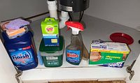 Photo showing the full cleaning supplies lot under the sink including dishwasher tabs, sponges, Windex spray, green container, and Ziploc bags.