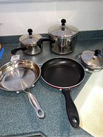 Photo showing three stainless steel pots with lids and two fry pans on kitchen countertop.