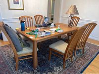 All six wooden dining chairs positioned around a wooden dining table with clutter on the table surface. The wood finish is warm-toned. Cushions are beige and appear lightly worn, but one cushion is discolored.
