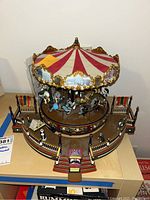 Electric carousel decoration with red and white striped canopy, gold decorative trim, miniature horses and figures attached on rotating platform, enclosed by detailed fencing and light posts, with a ticket booth and entry steps at front.