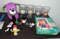 Lot overview showing purple plush dog, four Golden Girls glasses, Cat in the Hat figurines, Lego Santa figure, and boxed Golden Girls puzzle.