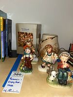 Photo of three Hummel figurines, empty Hummel box and beige vase with visible cracks