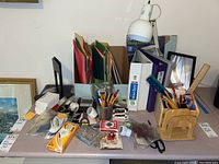 Wide view of lot showing file folders, binders, colored pencils, pens, desk lamp, clock, notebooks, and other office supplies arranged on a surface.