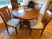 Round wooden dining table with pedestal base and four matching chairs with beige padded seats around it, placed on hardwood floor near window.
