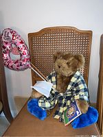 Full view of the teddy bear sitting on a wicker chair wearing a blue and cream checkered robe, holding a heart-patterned balloon and a face mask.