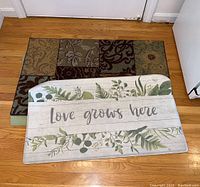 Two throw rugs, one with patchwork floral patterns and one with 'Love grows here' text and foliage design, both placed on hardwood floor.