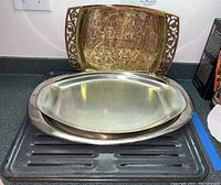 View of three trays stacked, showing the ornate embossed metal tray at the back, and two smooth metal trays including a roasting pan in front.