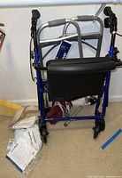 Folding walkers seen folded, including one Drive brand with black seat and blue frame along with additional health care aids and packaging.