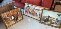 Three framed paintings of urban and architectural scenes, arranged on carpet. Gold ornate frames.
