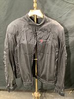 Front view of black Dainese vented riding jacket hanging on stand showing mesh ventilation panels and zip closure.