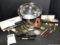 Array of mixed items on black background including oval silver-plated tray, watches, keychains, pens, knife pouch and small accessories