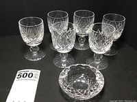 Seven piece set of Waterford crystal: six port wine glasses in Boyne pattern and one small ash receiver in Colleen pattern displayed on black background.