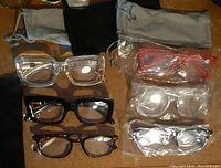 Six pairs of reading glasses displayed with three glasses bags in black and gray fabric. Three pairs unwrapped showing frame styles in clear, black, and tortoiseshell colors.