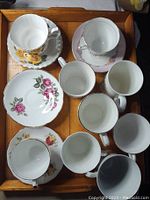Overview of nine china tea cups and seven saucers on wooden tray showing all cups and plates with floral designs