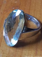 Close-up of silver ring with large oval green faceted stone on wooden surface showing reflective facets and polished band.