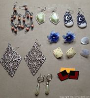 Photo showing nine pairs of assorted earrings with varied designs and colors on a beige background.