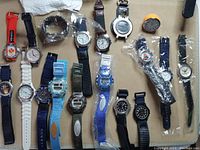 Overhead view of 17 assorted watches and one small timer device showing variety of band colors and types, most watches designed for wrist wear with analog or digital faces.