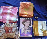 Four scarves arranged on blue fabric showing various patterns and colors, a gold-tone scarf pin, and spiral-bound scarf tying book.