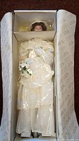 Top view of boxed bride doll named Flora in white lace bridal gown, with plastic wrapping and bouquet visible inside box