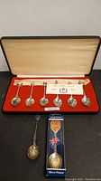 Full display box showing 6 silver-plated spoons, red velvet lining, certificate or informational paper behind spoons, plus two additional separate collectible spoons.