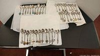 Photo of approx 50 silver-tone collector spoons arranged on white cloths showing various enamel and sculpted handle emblems and designs.