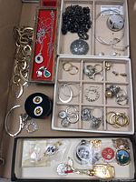 Top down view showing mixed costume jewelry including chain link bracelet, silver-tone necklaces with pendants, black beaded necklace with ornamental pendant, silver-tone circular choker, and compartments with various earrings.