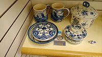 Top-down view of the lot showing all items grouped together on a shelf: two large mugs decorated with Dutch scenes, three lidded serving dishes with blue floral patterns, and a small decorative clog figurine.
