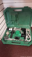 Open green carrying case showing Hitachi rotary hammer drill with attached power cord, auxiliary handle, and accessories arranged inside.