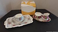 Royal Albert tea set with cup, saucer, cream, sugar, and two trays arranged on a table with white and yellow box in background