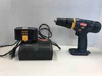 Ryobi cordless drill, battery pack, and charger with cords arranged side by side on a white surface and backdrop.