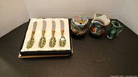 Photo of four butter utensils with leaf handle design in cushioned box, and sugar bowl with lid, two small creamer jugs with multicoloured floral pottery glaze