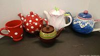 All four teapots and the coordinating mug displayed together on a black surface against a white wall.