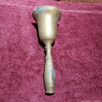 Full view of the old brass hand bell laying on a purple fabric showing cylindrical brass bell and worn wooden handle.