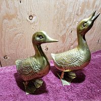 Two brass duck figurines side by side, showing front and side views, highlighting embossed feather detailing and different beak positions.