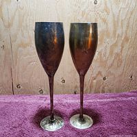 Front view of two vintage silver champagne flutes standing upright on a purple cloth with visible tarnish and oxidation on surfaces.