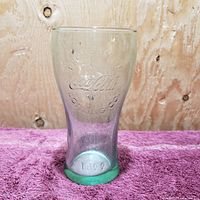 Close-up view of clear Coca Cola glass with embossed logo, text 'Property of Coca Cola Bottle Co' and '1899' year on the base, placed on purple fabric with wooden background.