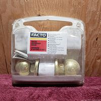 Front view of Facto keyed entrance knob set in clear plastic case showing two brass finish knobs, mounting hardware, and instruction/key packet inside.