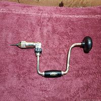 Fuller ratcheting hand drill laid flat on a burgundy textured cloth with drill bit inserted, showing side view of crank handle and ratchet mechanism.