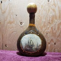 Front view of rounded vintage glass liquor bottle covered in brown leather with hand-painted sailing ship scene and wooden round cork stopper, set against wooden backdrop and purple towel surface.