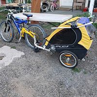 Side view of the Burley Tail Wagon pet trailer attached to a blue mountain bike on gravel surface, showing the frame, wheels, and hitch mechanism.