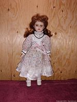Front full-body photo of vintage porcelain doll against a wooden background showing the doll's brown curly hair, floral print dress with lace and pink ribbon, white shoes.
