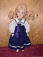 Front view of vintage porcelain doll with blonde curly hair and blue dress against wood backdrop.