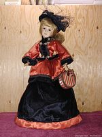 Photo of vintage porcelain doll standing against a light wooden wall, dressed in orange satin and black velvet with veil hat and holding a striped handbag.
