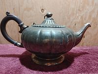 Front-left side view of vintage silver plated copper tea pot showing fluted body and wooden handle on red cloth background.