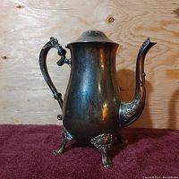 Side view of vintage silver plated tea pot showing ornate handle, spout, and feet against a wooden background on maroon fabric.